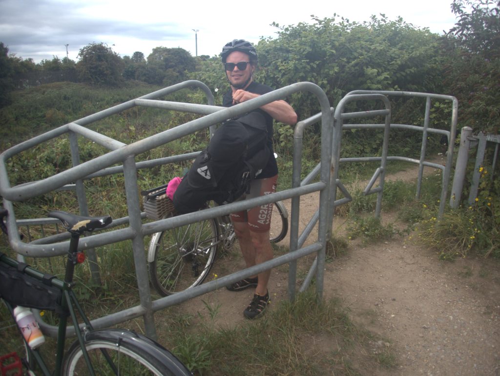 Photo of someone stuck behind a steel barrier, supposedly to stop motorbike trespassers. They are handing over their pannier bags before they lift their bike over it.