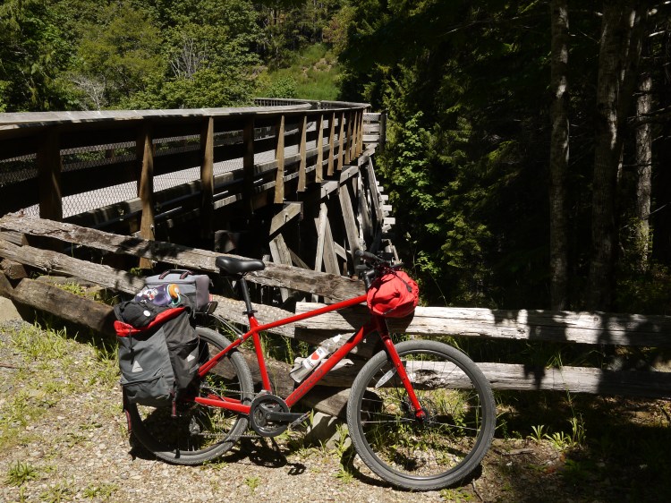 Bicycle learning against a fence. It's laden with bags and a large trestle bridge is in the background.